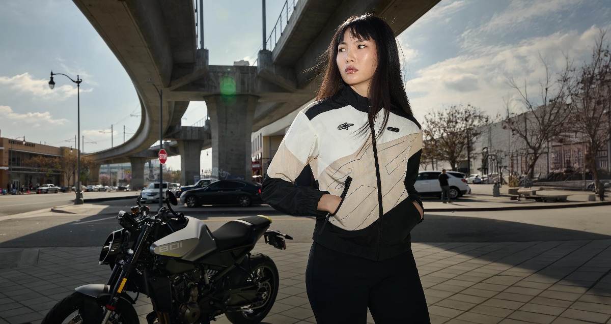 asian girl using an Alpinestars chaquet under a bridge in a city with a motorbike on the right side